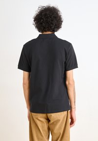 Black short-sleeve polo shirt with a pointed collar, worn with tan corduroy pants. Smooth texture, minimal design, no visible patterns.