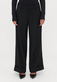 Melni platleņķu bikses ar gludu tekstūru, salocītām piedurknēm un pieguļošu jostu. Nēsātas ar norādītiem melniem zābakiem.