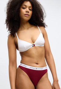 Balta trīsstūru matu un plānu siksniņu krūšturis ar nelielu logotipa detaļu; bordo krāsas tanga ar balto logotipa jostu. Gluda auduma tekstūra.