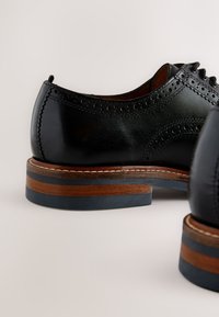 Black leather shoes with brogue detailing, rounded toe, and stacked wooden heel with contrasting blue accent. Smooth texture and classic design.