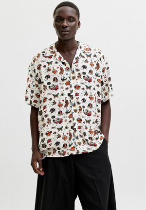 Osoba o ciemnej karnacji ubrana w białą, krótkorękawową koszulę z kolorowymi wzorami w stylu tatuażu i luźne czarne spodnie, stojąca na jednolitym tle.