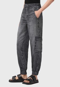 Schwarze Denim-Cargohosen mit elastischen Bündchen, Seitentaschen und einer strukturierten Oberfläche. Kombiniert mit schwarzen Pantoletten mit doppelten Schnallen.