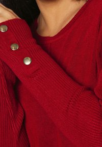 Pull rouge côtelé avec des accents de boutons dorés sur le poignet. Texture douce avec une coupe ajustée et un col rond.