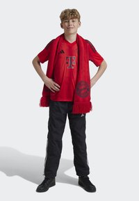 Maillot rouge du FC Bayern Munich avec des accents noirs, une écharpe rouge à franges, un pantalon cargo noir avec des rayures blanches et des chaussures de sport noires.