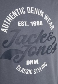 Grått tyg med vit och mörkblå text: "Äkta Denim Wear Grundat 1990 Jack & Jones DNM. Klassisk Stil."