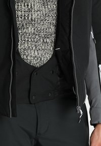 Schwarze, mit Reißverschluss versehene Jacke über einem geknöpften schwarzen Weste und einem schwarz-weißen Strickpullover, kombiniert mit schwarzen Hosen und sichtbar grauen Hemdsärmeln.
