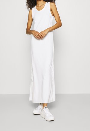 Woman wearing a sleeveless white maxi dress paired with white platform sneakers, standing against a plain light background.