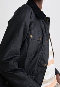 Černá bunda s tmavým kordovým límcem, s velkými předními kapsami a mosaznými zaklapávacími knoflíky, nošená přes pruhovanou košili.