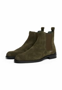 Par de botines Chelsea de ante verde oliva con paneles laterales elásticos y suelas negras sobre un fondo blanco.