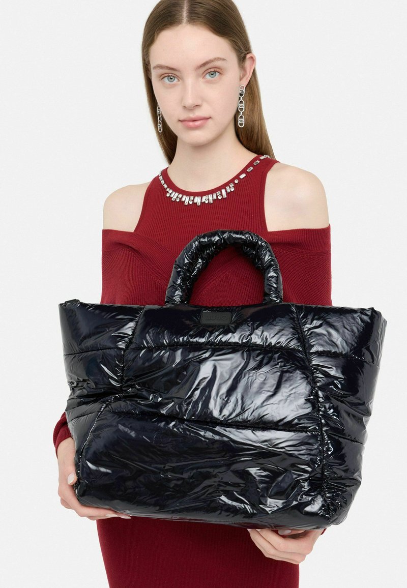Borsa a tote nera in stile piumino con finitura lucida, design trapuntato e manici corti. Presenta un logo visibile sul fronte. Ha una forma arrotondata.