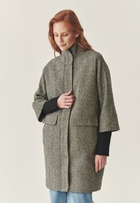 Cappotto in misto lana grigio con colletto alto, ampie tasche frontali e maniche a tre quarti. Tessuto testurizzato con un motivo sottile.