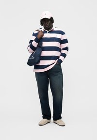 Mann iført rugbyskjorte med rosa og marineblå striper, mørke jeans, beige sko, rosa caps, og bærer en marineblå handlenettveske over den ene skulderen.