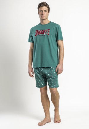 Joven con pantalones cortos verdes con motivos de Snoopy y camiseta a juego, de pie descalzo contra un fondo blanco.