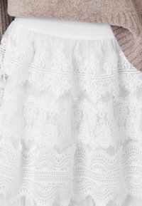 Jupe en dentelle blanche avec des motifs floraux et géométriques complexes associée à un pull beige en tricot.