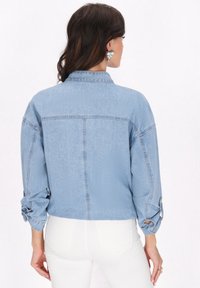 Jasnoniebieska dżinsowa kurtka z kołnierzykiem, zapinanymi mankietami i luźnym krojem, z subtelną fakturą i detalem w postaci szwu na plecach.