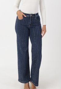 Vida jeans i denim med vertikala silverränder, hög midja, framfickor och knäppning med knappar. Mörkblå färg och slät textur.