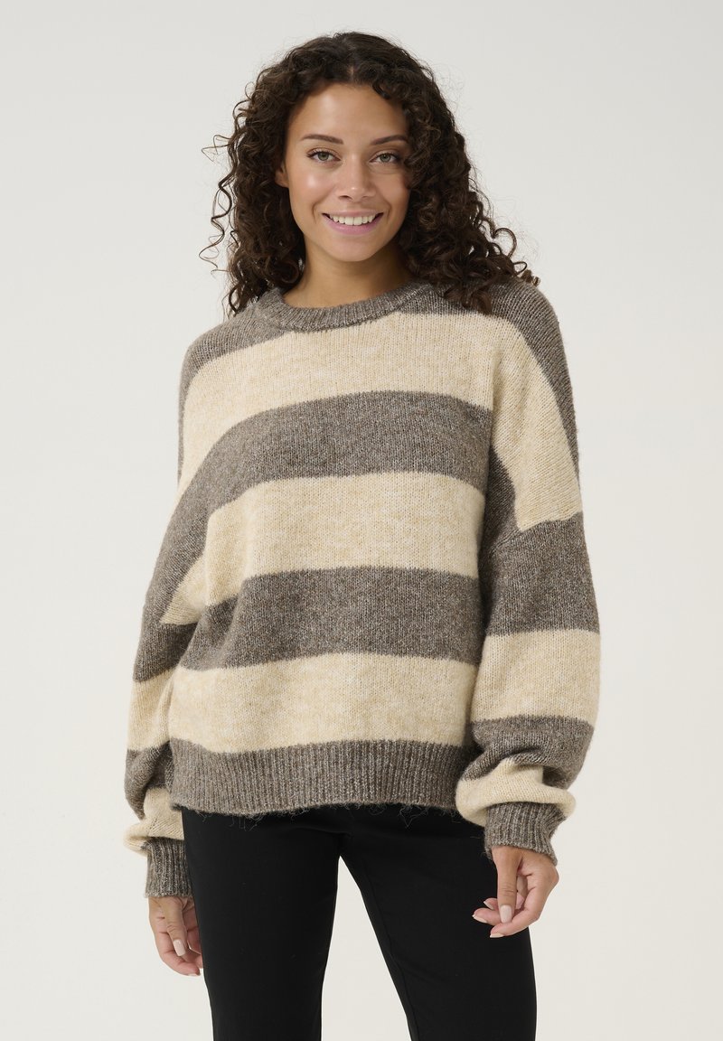 Prugasta pulover s oversize krojem, ki ima bež in rjave stripe, rebraste manšete, mehak material in okroglo ovratnico. Usklajen z črnimi hlačami.