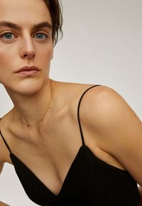 Vestido negro de tirantes finos con un escote en V profundo; tejido suave; collar delicado dorado con pequeños acentos alrededor del cuello.