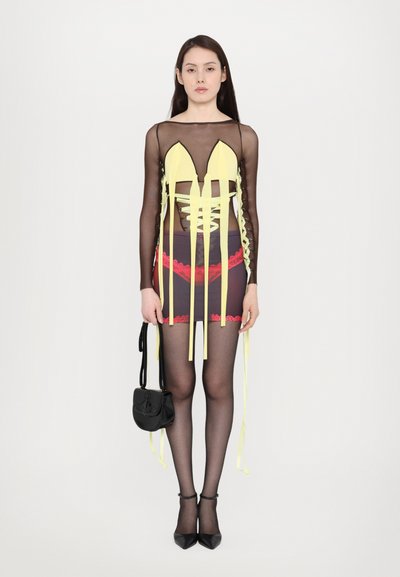 Durchsichtiges schwarzes langärmliges Kleid mit gelben Akzenten, Schnürung an der Vorderseite und rotem Spitze am Saum, kombiniert mit schwarzen Riemchen-Absätzen und einer kleinen Tasche.