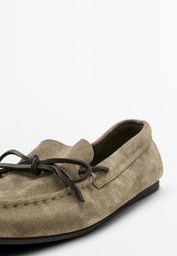 Wildleder-Slipper in Olivgrün, mit runder Zehe, gesteppten Details und einem dunkelbraunen Schnürsenkelbogen-Akzent an der Vorderseite. Flache Sohle.