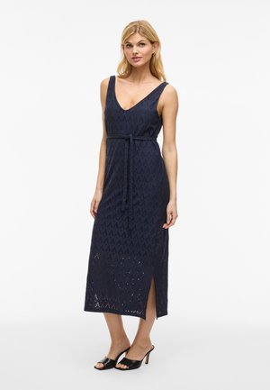 Robe maxi bleu marine avec un décolleté en V, sans manches, taille ceinturée, motif à découpes en losange et fentes latérales, associée à des escarpins noirs.