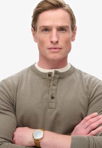 Lichtbruine henley-shirt met drie knopen, gestructureerde stof, gelaagd over een crèmekleurige kraag. Draagt een gouden horloge met een bruine leren band.