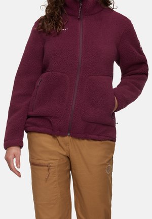 Femme portant une veste polaire bordeaux zippée et un pantalon marron clair avec une poche zippée sur la cuisse, debout avec une main dans la poche.