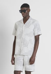 Camisa de manga corta gris claro y pantalones cortos a juego con un patrón de hoja de helecho. Llevado con gafas de sol rectangulares negras.