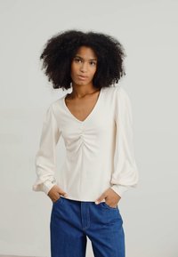 Light beige long-sleeve blouse with a V-neck and gathered detailing at the chest, paired with blue denim jeans. Smooth texture and relaxed fit.