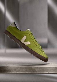 Zelené semišové tenisky s texturovaným povrchem, bílým logem a kontrastní hnědou gumovou podrážkou, se šněrováním a kulatou špičkou.