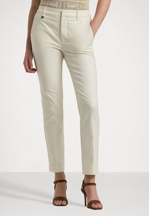 Personne portant un pantalon beige slim et des sandales marron à talons hauts, debout devant un fond uni.