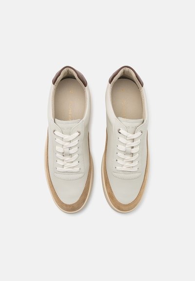 Et par hvide lædersneakers med beige ruskinds tåspidser, hvide snørebånd og mørkebrun hæl-accenter på en ensfarvet lys baggrund.