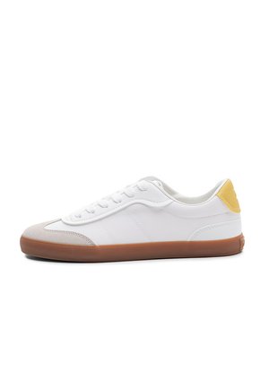 Biały niski sneaker z gumową podeszwą, beżowym czubkiem, białymi sznurowadłami i żółtą pętlą na pięcie, pokazany z profilu na białym tle.