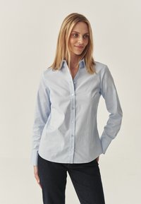 Camicia azzurro chiaro con colletto, maniche lunghe e una texture liscia. Presenta un orlo arrotondato e bottoni scuri. Indossata con jeans scuri.