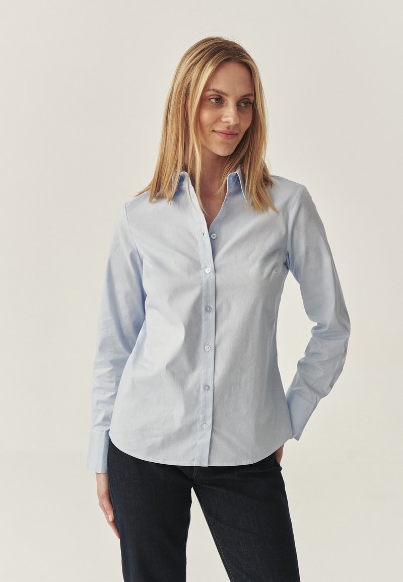 Camicia azzurro chiaro con colletto, maniche lunghe e una texture liscia. Presenta un orlo arrotondato e bottoni scuri. Indossata con jeans scuri.