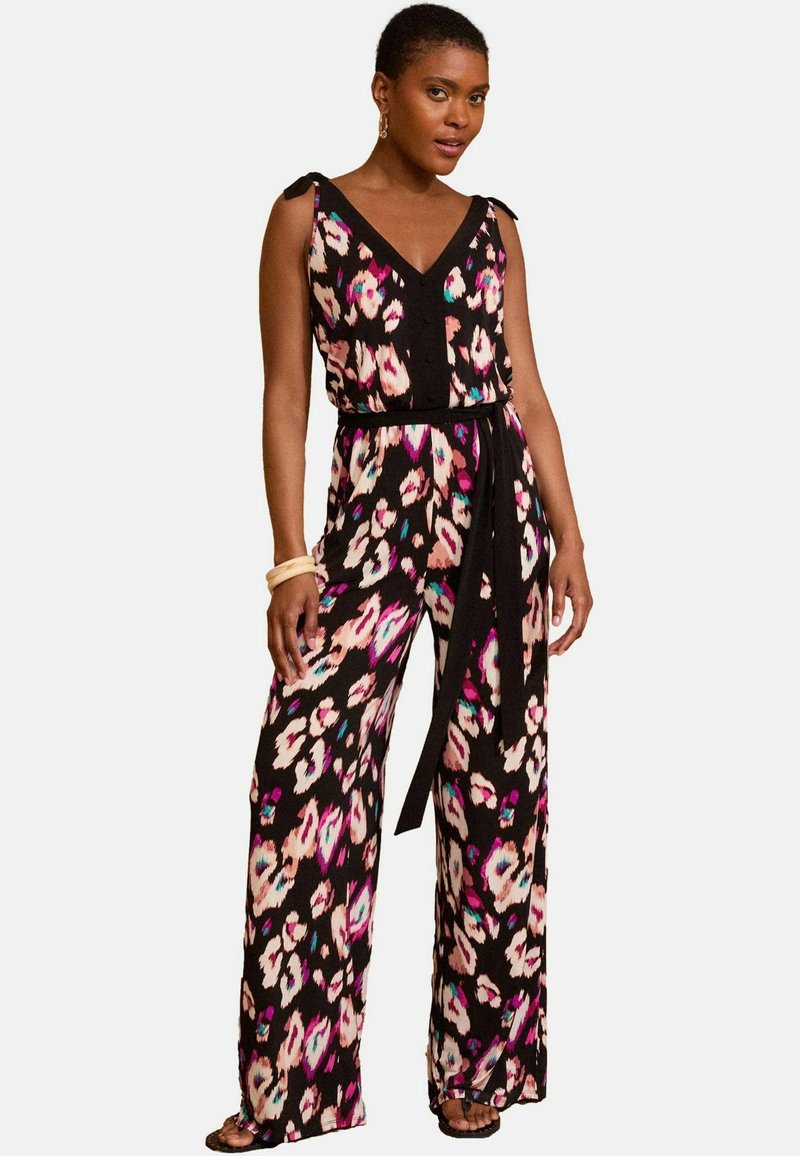 Ženska v brezrokavnem širokem kombinezonu s črno, roza, belo in modro cvetličnim vzorcem ter zavezanim pasom.