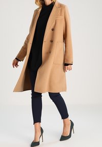 Abrigo de mezcla de lana color camello con un diseño estructurado, frontal de doble botonadura, dos grandes bolsillos y un ajuste entallado, combinado con pantalones azul marino.