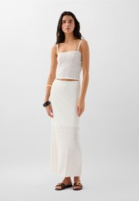 Witte gebreide crop top met dunne bandjes, gecombineerd met een lange, witte, textuurrok met patroon details in het breiwerk. Bruine sandalen maken de look compleet.
