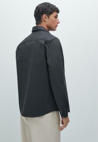 Black long-sleeve shirt with a standard collar, smooth texture, and buttoned cuffs, viewed from the back, paired with cream trousers.