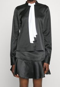 Blusa di raso nera con un nastro bianco al collo, con colletto alto e maniche lunghe, abbinata a una gonna svasata di raso coordinata.