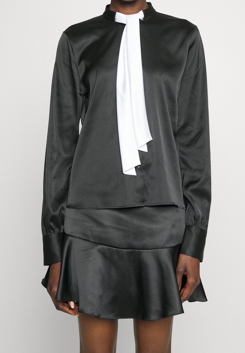 Blusa di raso nera con un nastro bianco al collo, con colletto alto e maniche lunghe, abbinata a una gonna svasata di raso coordinata.