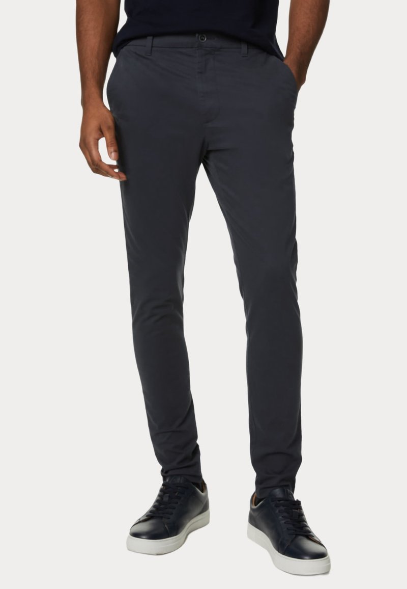 Homme portant un pantalon slim gris foncé et des baskets noires à semelles blanches, debout les mains dans les poches devant un fond uni.