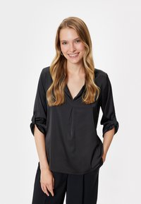 Zwarte blouse met lange mouwen, een V-hals en plooidetail. Beschikt over opgerolde mouwen en een gladde, satijnachtige textuur.