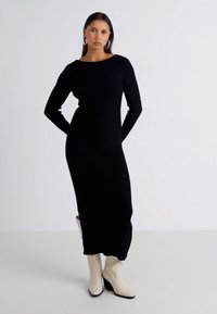 Robe noire en tricot côtelé avec des manches longues, un col rond et une silhouette ajustée. Accompagnée de bottes à talons carrés de couleur crème.
