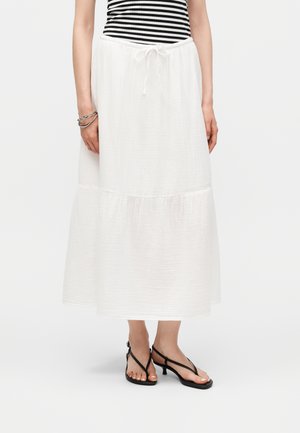 Frau trägt einen weißen gestuften Midirock mit Tunnelzug in der Taille, ein schwarz-weiß gestreiftes Oberteil, silberne Armbänder und schwarze Sandalen mit Absatz.