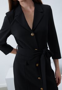 Kvinna iklädd en svart blazerkjol med bälte och guldknappar, trekvartslånga ärmar och ett knytband i midjan, stående mot en enfärgad bakgrund.