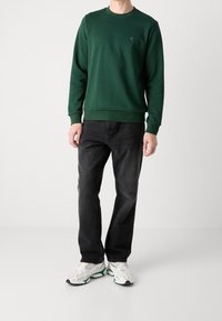 Felpa verde scuro con scollo rotondo, polsini e orlo a costine. Abbinata a jeans neri e sneakers colorate, con un design testurizzato.