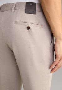 Beige texturerade byxor med en bakficka som har en brun knapp, samt en mörk läderetikett med "JOOP!" präglat på midjebandet.