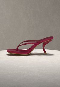 Κόκκινο πέδιλο με ψηλό τακούνι, λεπτά διπλά λουράκια και ανοιχτό δάχτυλο, τοποθετημένο σε ένα ελαφρύ στρογγυλό βάθρο σε ουδέτερο φόντο.