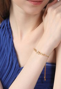 Gold bracelet featuring floral designs with white and clear stones. The band is delicate, with a chain extension and two small beads.